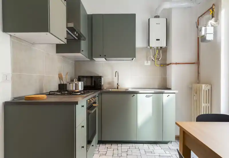 Cuisine compacte et moderne avec meubles vert mat, four encastré et plaque à gaz, évier en inox et petite table en bois — lumineuse et bien mise en scène pour une annonce locative.