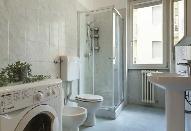 Salle de bain lumineuse et propre avec cabine de douche en verre, WC, bidet, lavabo et une machine à laver près d'une fenêtre apportant la lumière naturelle.