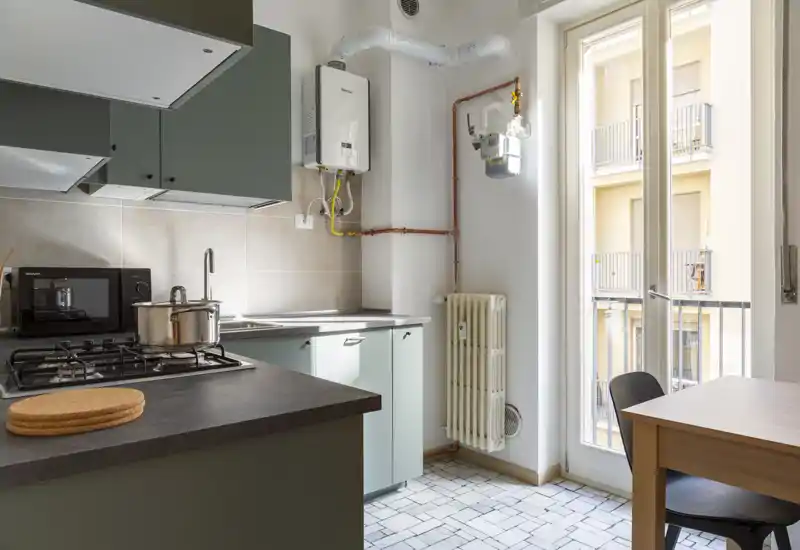 Cuisine compacte et moderne avec placards verts, cuisinière à gaz, micro-ondes et évier ; lumineuse grâce à une grande porte-fenêtre donnant sur le balcon.