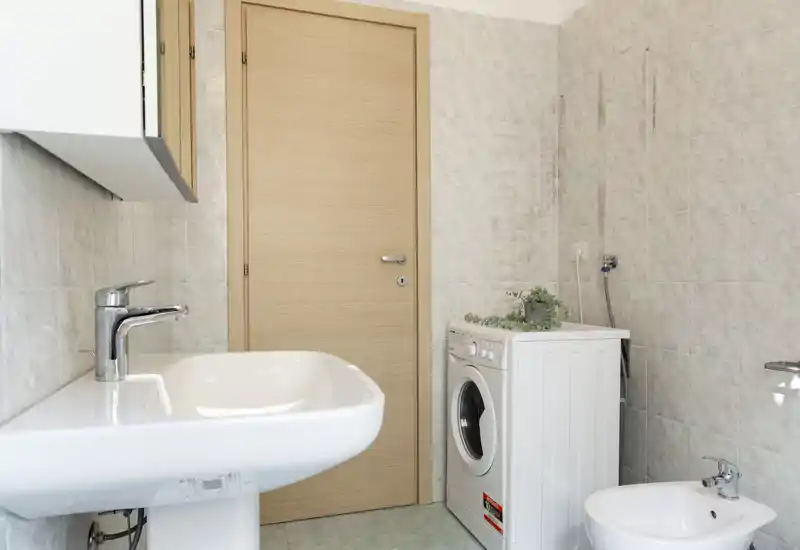 Salle de bains propre et lumineuse avec lavabo sur colonne, bidet et lave-linge frontal près de la porte en bois. Carrelage clair et équipements simples pour un espace fonctionnel.