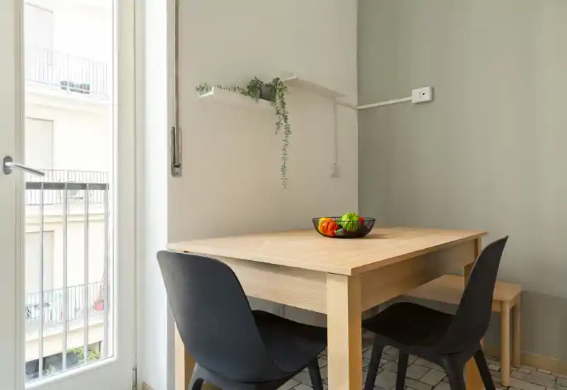 Coin repas compact avec une table en bois clair, deux chaises noires et un bol de fruits décoratif, éclairé par la lumière naturelle de la porte du balcon — petit espace repas lumineux et moderne.