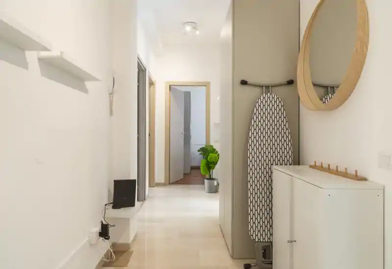 Entrée/couloir lumineux et minimaliste aux tons neutres, miroir mural, meuble de rangement et planche à repasser rangée proprement — bien éclairé et soigné.