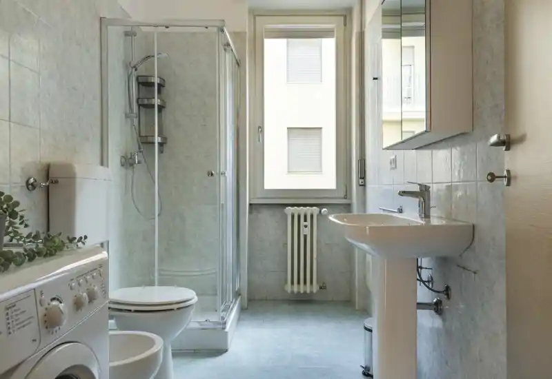 Salle de bain moderne et lumineuse avec cabine de douche en verre, lavabo colonne, toilettes et machine à laver à côté du bidet. Carrelage neutre et fenêtre offrant une belle lumière naturelle, espace propre et fonctionnel.