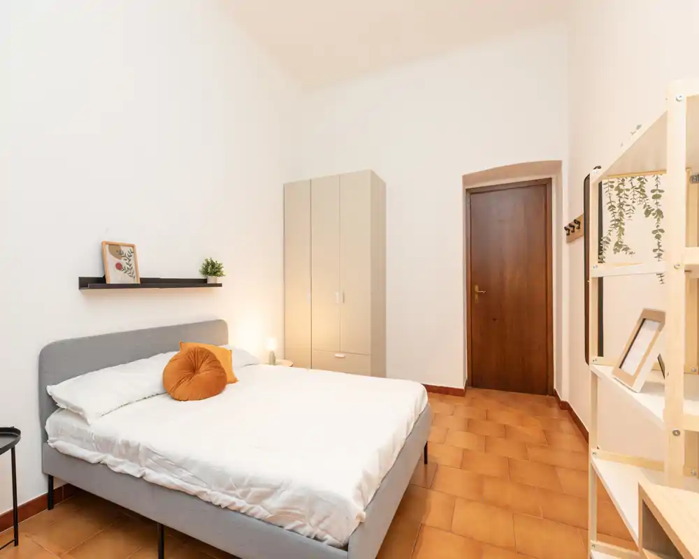 Bright, minimalist bedroom with a double bed, warm terracotta floor and a standing wardrobe. The room is simply decorated with a round cushion and a small shelf above the headboard, creating a calm, modern atmosphere.