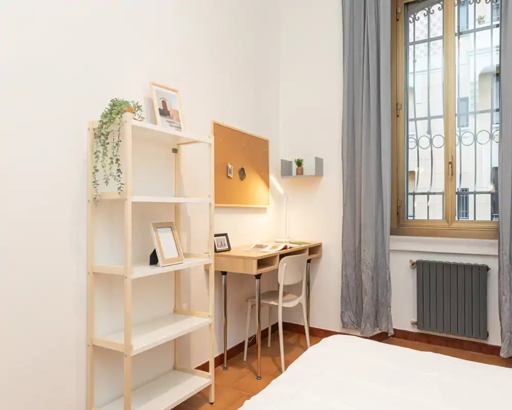 Bright, simply styled bedroom corner with a visible bed, a small wooden desk with lamp and chair, open shelving and a large window letting in natural light.