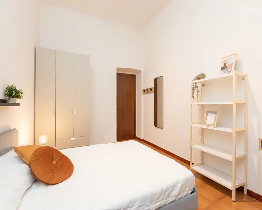 Bright, minimalist bedroom with a large bed, warm accent cushion and practical storage including a wardrobe and shelving unit.