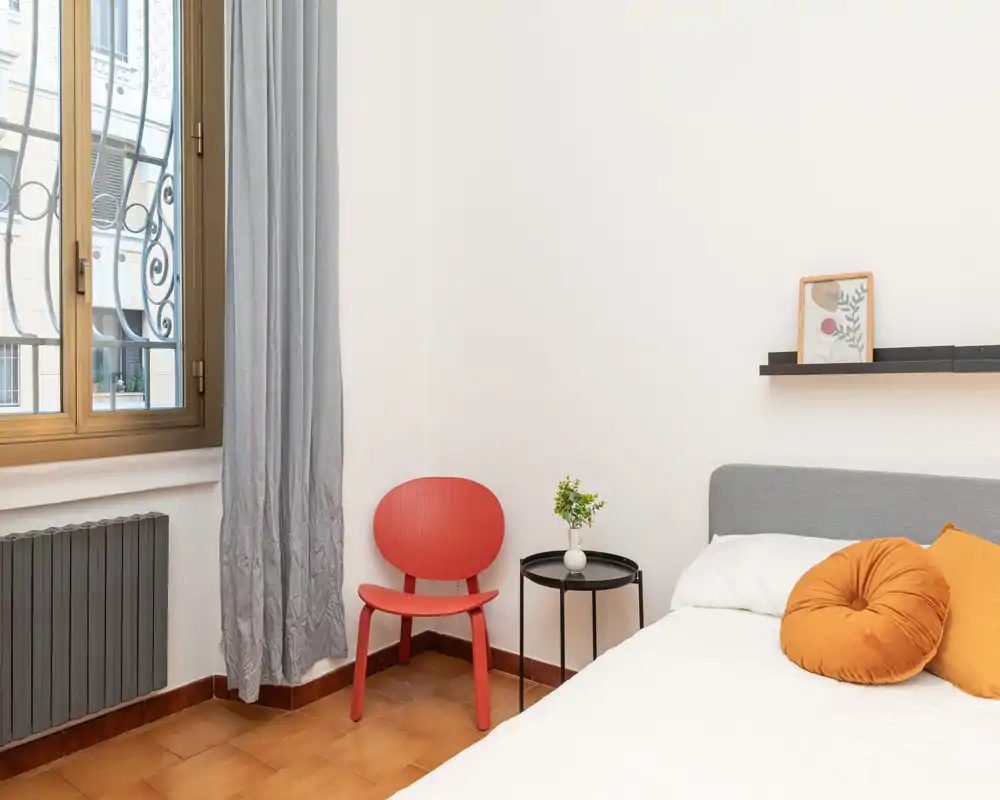 Bright, modern bedroom corner with a visible bed head, warm cushions, a small bedside table and a red accent chair by the window — cozy and well styled for a rental listing.