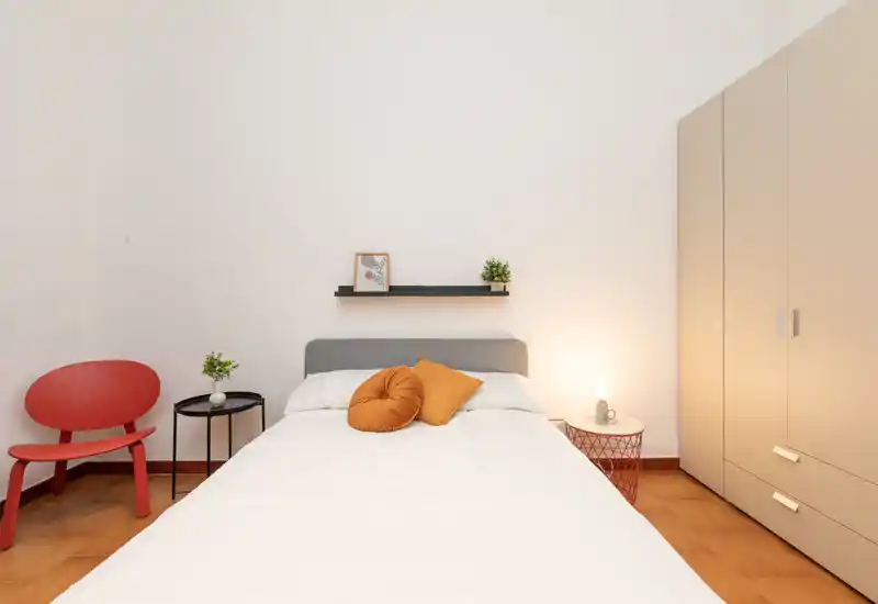 Bright, modern bedroom with a neatly made double bed, warm accent cushions and bedside lighting; simple wardrobe and tasteful minimalist decor.