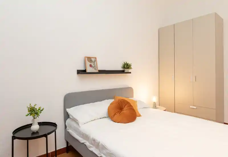 Bright, neatly styled bedroom with a grey upholstered bed, warm accent pillows and a tall wardrobe. Small bedside lamp and plants add a cozy touch.