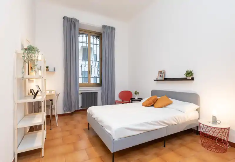 Bright, modern bedroom with a comfortable double bed, warm terracotta floor and simple, stylish furnishings including a small desk and shelving.
