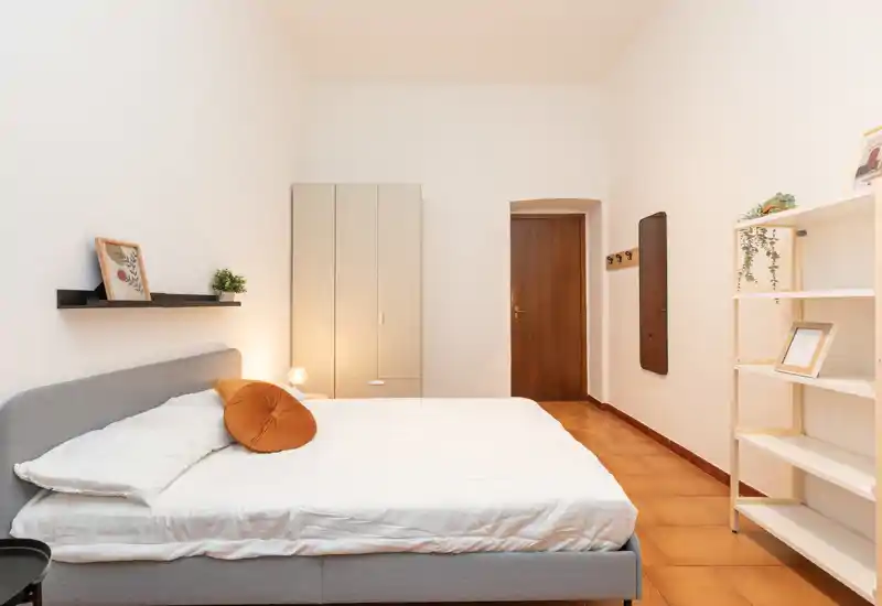 Bright, minimalist bedroom with a large double bed, soft linens and warm accents. Neutral tones, practical wardrobe and shelving create a calm, inviting space ideal for restful stays.