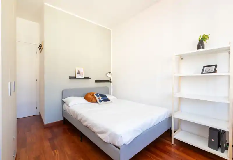 Bright, minimal bedroom with a neatly made double bed, warm wooden floors and simple shelving — ideal for listing photos highlighting a cozy, modern single/double room.