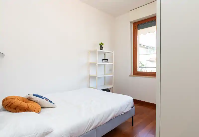 Bright, minimalist bedroom with a neatly made double bed, wooden floor and a window providing natural light. Simple shelving and soft decorative cushions create a calm, welcoming atmosphere.