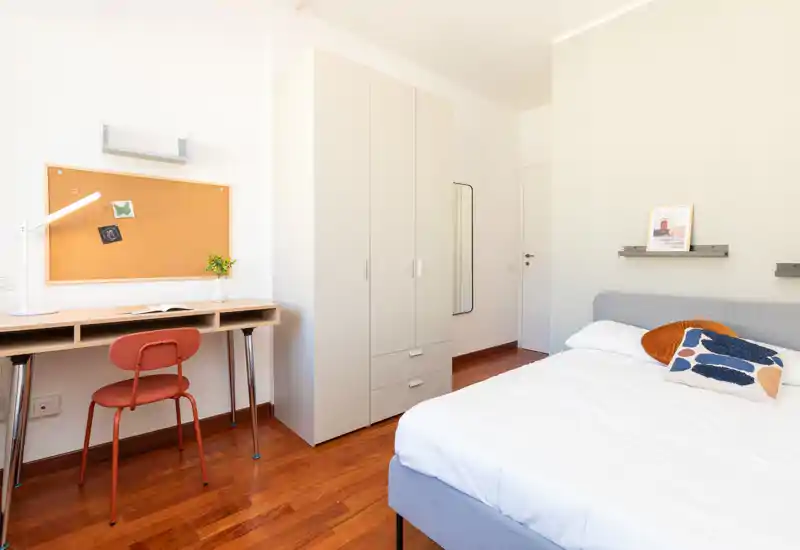 Bright, modern bedroom with a comfortable bed, decorative cushions and a study desk beside a cork board; warm wooden floors and clean minimalist storage.