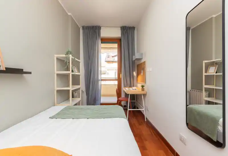 Bright, neatly staged single bedroom with a comfortable bed, desk and a balcony door that provides natural light; mirror on the wall adds depth and a shelving unit offers storage.