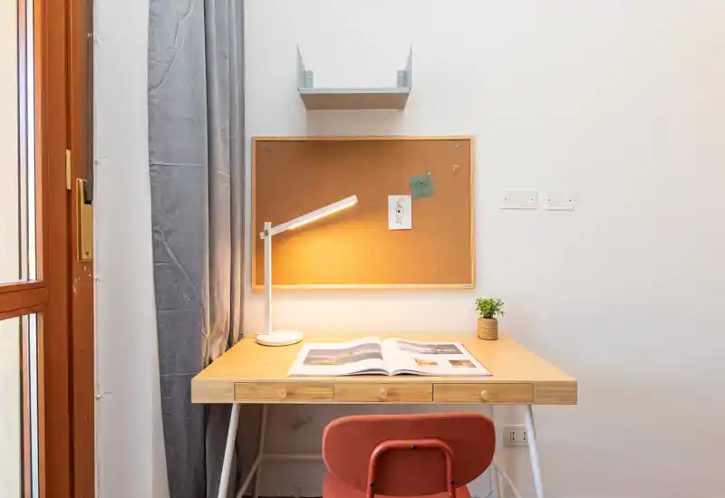 Compact, well-lit workspace with a wooden desk, adjustable desk lamp, cork notice board and a comfortable chair — ideal for studying or remote work.