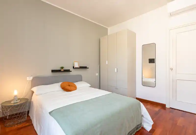 Chambre lumineuse et moderne avec lit double bien fait, lampe de chevet, armoire et miroir intégral — tons neutres et sol en bois chaleureux.