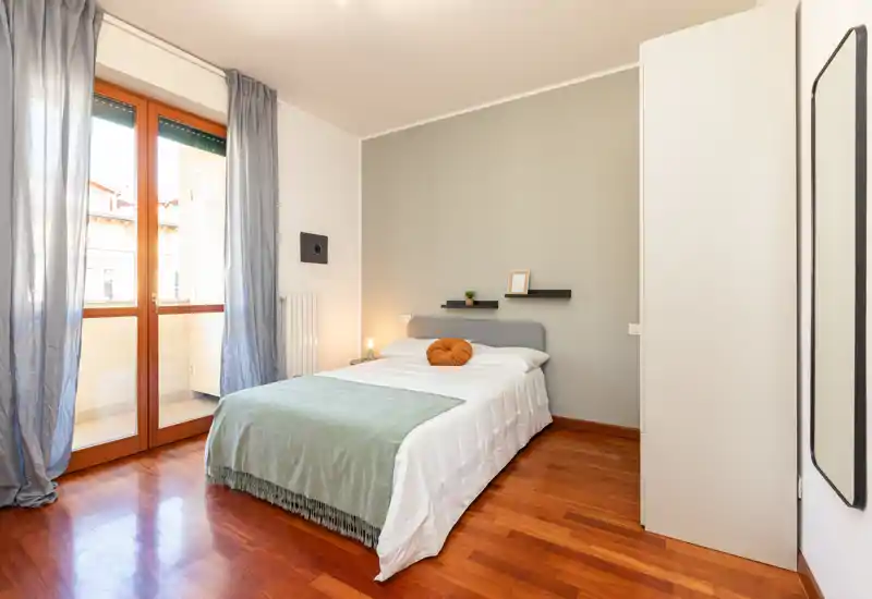 Chambre lumineuse et moderne avec un lit double habillé de linge blanc, un plaid vert et un coussin rond décoratif. Grande porte-fenêtre apportant beaucoup de lumière naturelle ; étagères minimalistes et parquet chaleureux pour une atmosphère cosy et contemporaine.