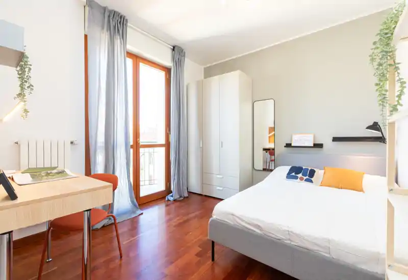 Chambre lumineuse et moderne avec lit double, armoire, bureau et grande fenêtre laissant entrer une belle lumière naturelle. Parquet chaleureux, textiles doux et touches décoratives simples pour une ambiance accueillante.