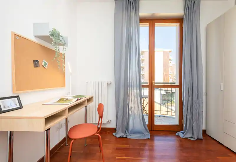 Coin de chambre lumineux et soigné avec bureau en bois, panneau liège et porte-fenêtre laissant entrer la lumière naturelle ; armoire visible sur le côté.