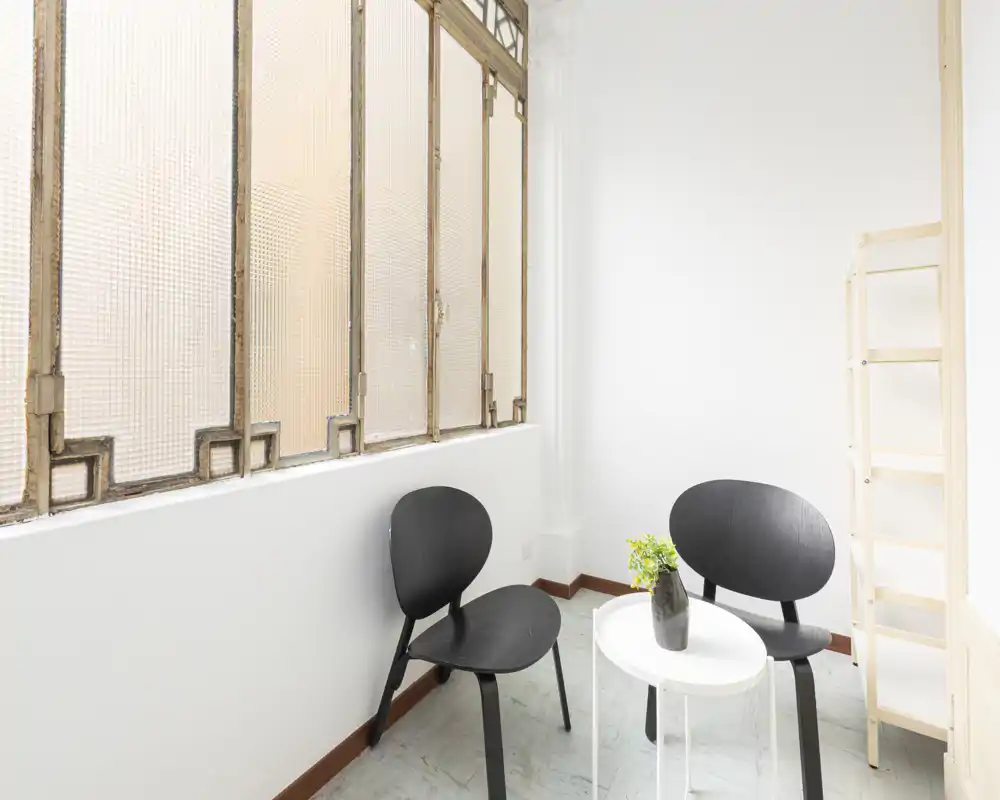 Angolo conversazione minimal e luminoso con due sedie nere moderne, un piccolo tavolino bianco e un vaso decorativo vicino a grandi finestre strutturate — perfetto per leggere o aspettare.