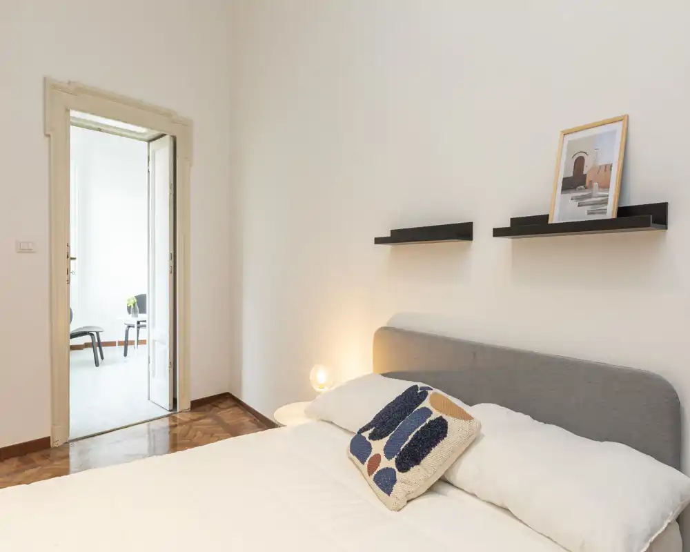 Camera da letto elegante con testiera imbottita grigia, biancheria bianca e cuscino decorativo; lampada da comodino e mensole minimal creano un'atmosfera accogliente e moderna.