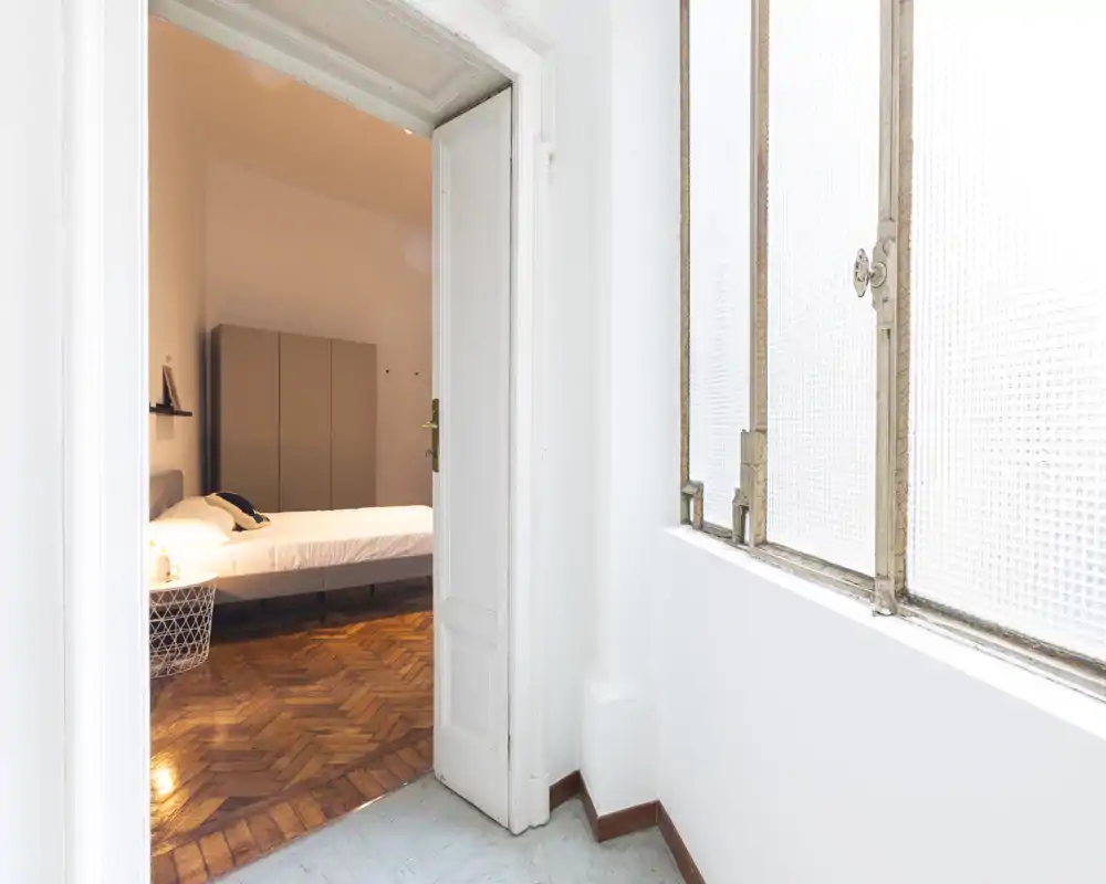 Camera vede una camera da letto luminosa attraverso una porta aperta: letto perfettamente rifatto, pavimento in parquet a spina di pesce e armadio sullo sfondo — ideale per annunci di affitto.