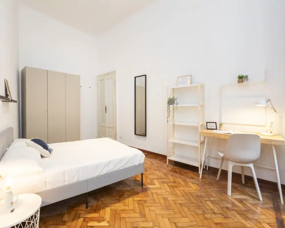 Camera da letto luminosa e minimal con comodo letto matrimoniale, armadio, scrivania e scaffale; parquet in legno caldo e buona illuminazione naturale e da lampada creano un ambiente accogliente.
