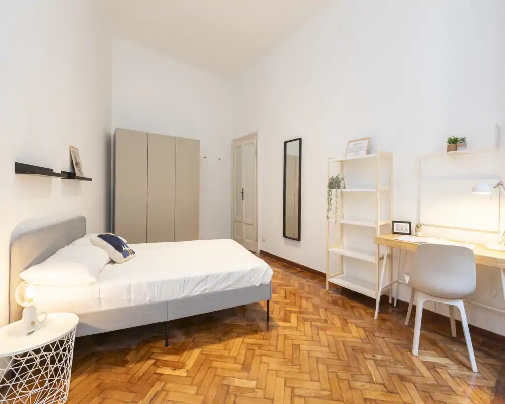 Camera da letto luminosa e moderna con ampio letto matrimoniale, pavimento in parquet, armadio e una postazione di lavoro ordinata — ideale per soggiorni confortevoli e lavoro produttivo.