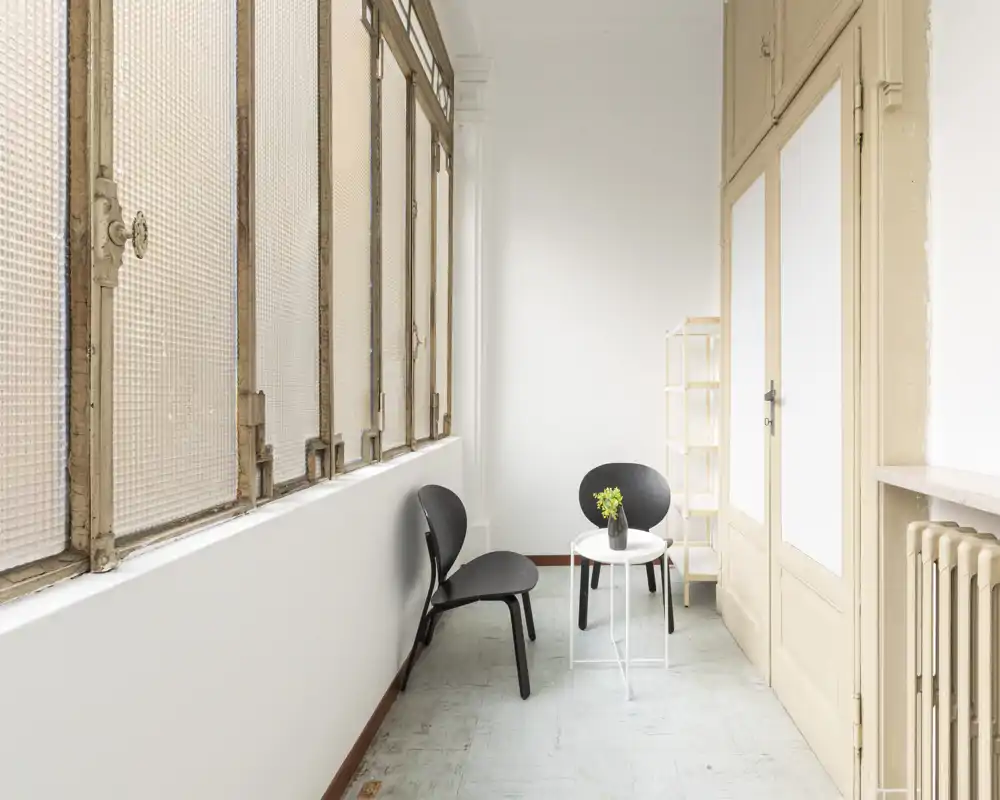 Piccolo ingresso o corridoio luminoso con ampie finestre strutturate, due sedie nere moderne e un piccolo tavolino rotondo — area di transizione pulita e minimalista.