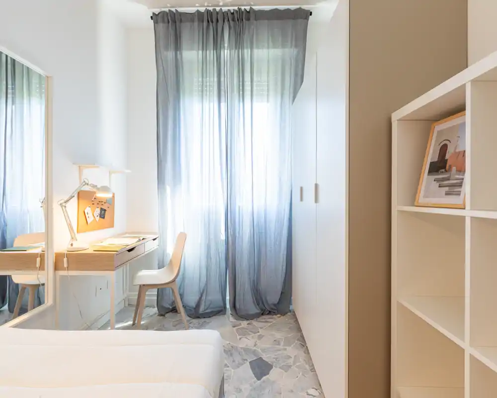 Camera da letto luminosa e dallo stile morbido con letto visibile, scrivania e armadio; tende leggere filtrano la luce naturale per un'atmosfera tranquilla.