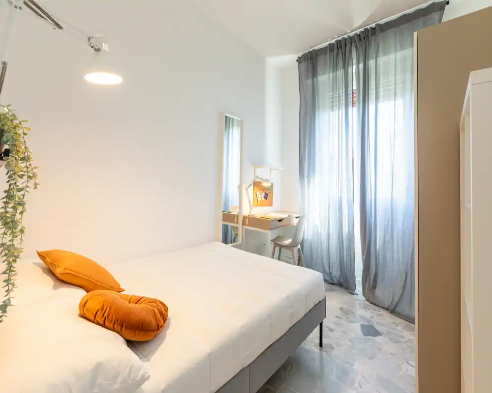 Camera da letto moderna e luminosa con letto matrimoniale, cuscini d'accento caldi, scrivania e finestra alta con tende leggere che diffondono la luce naturale.