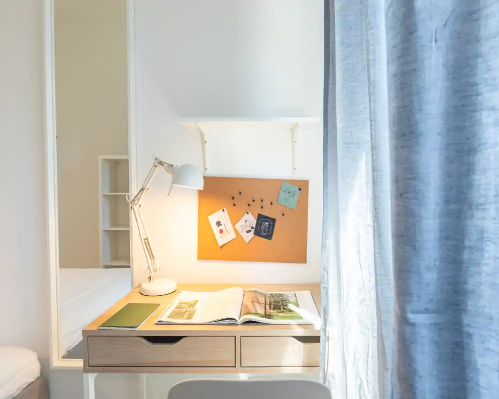 Angolo camera luminoso e minimal con una piccola scrivania, lampada e il letto parzialmente visibile a sinistra. La luce naturale filtra dalla tenda creando un accogliente angolo studio.