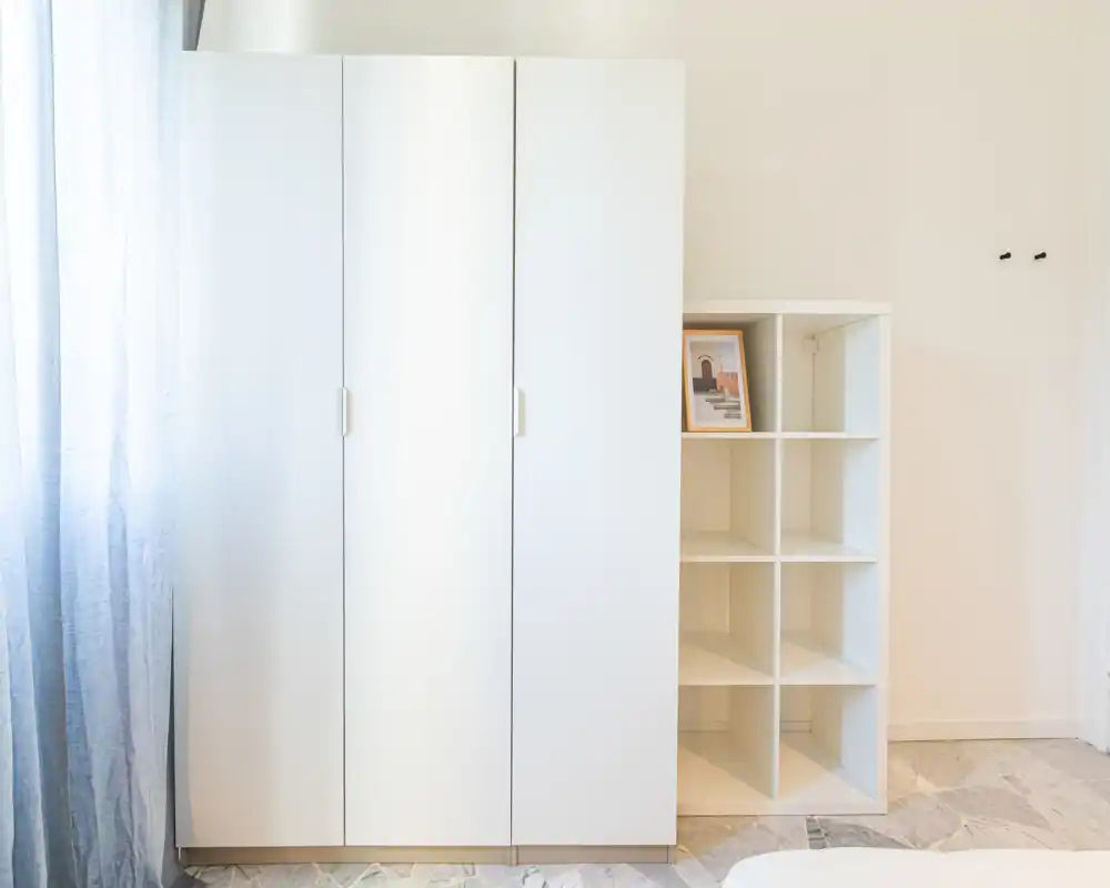 Angolo notte luminoso e minimale con un armadio bianco a tre ante e un modulo a cubi accanto a una finestra con tenda leggera, linee pulite e toni neutri.