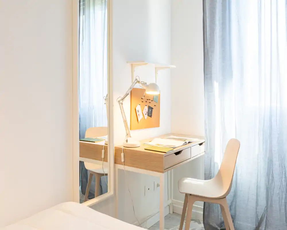 Angolo camera luminoso e minimale con scrittoio, lampada da lavoro e specchio a figura intera che riflette parte della stanza; tende leggere filtrano la luce naturale.