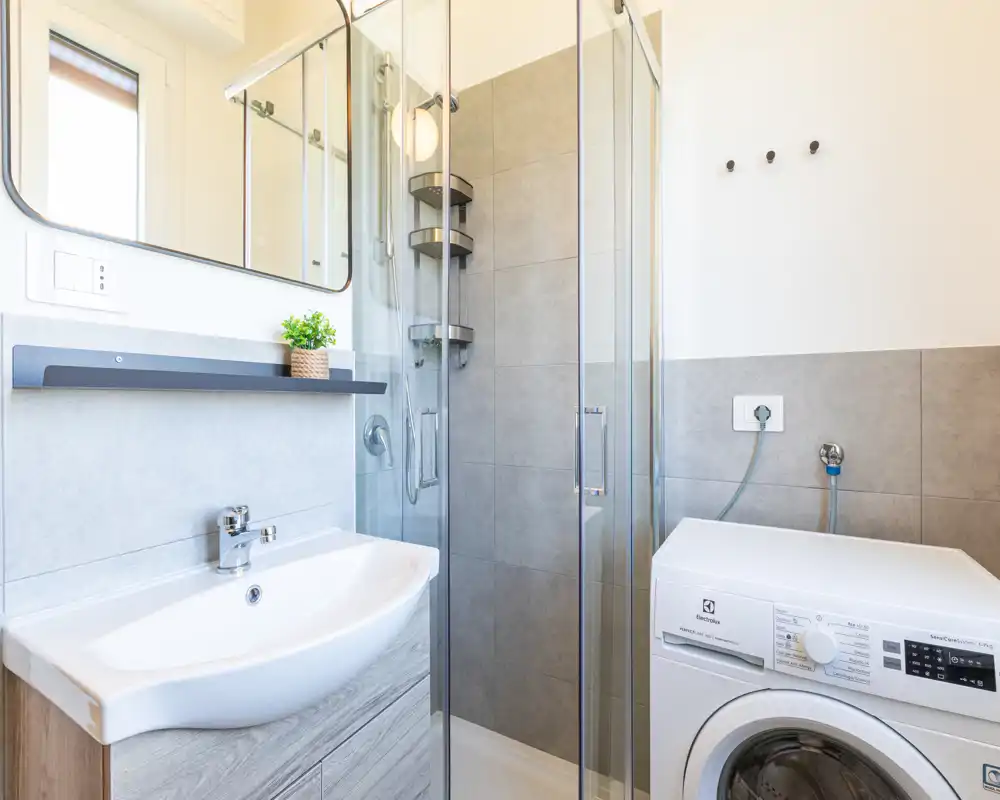 Bagno moderno e luminoso con box doccia in vetro, specchio a parete, lavabo con mobile e una lavatrice posizionata accanto alla doccia.