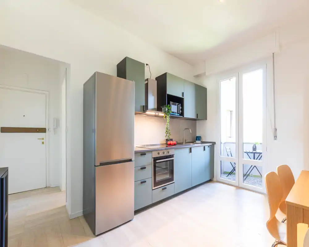 Cucina moderna a vista con elettrodomestici in acciaio inox, ante lisce e una porta sul balcone che illumina l'ambiente con luce naturale.