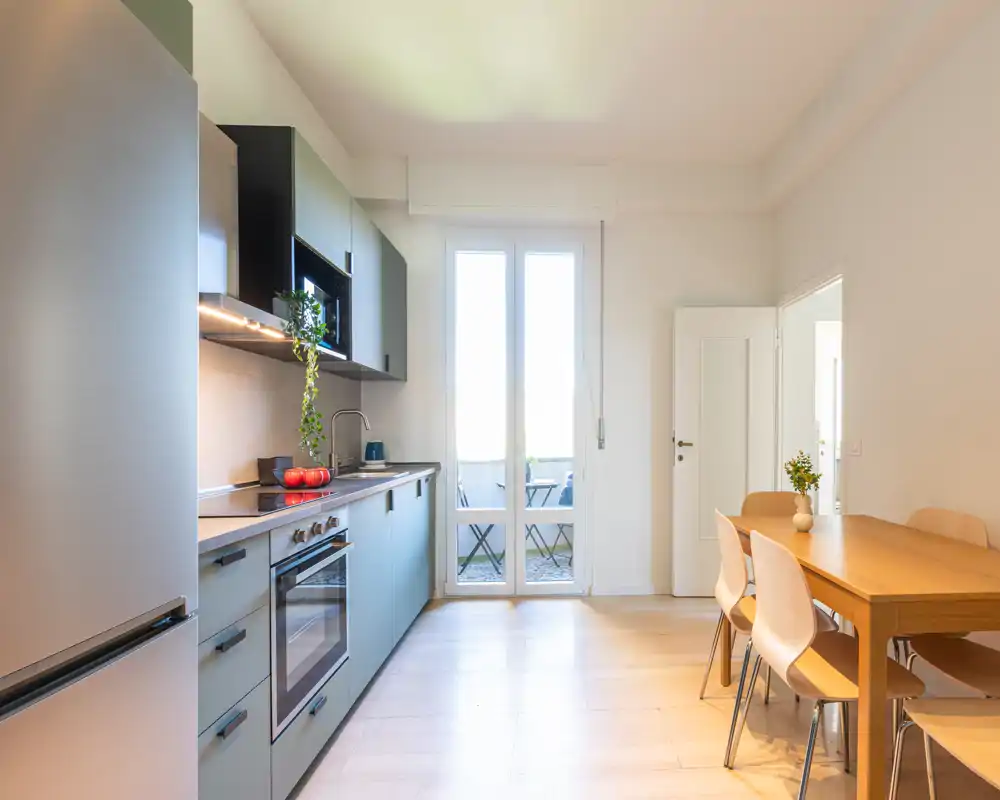 Cucina moderna su misura con elettrodomestici integrati, tavolo da pranzo in legno e sedie, illuminata da luce naturale proveniente dalla porta del balcone — perfetta per annunci di appartamenti.