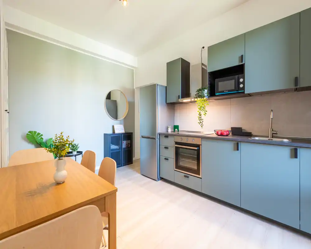 Cucina moderna e luminosa con ante blu opache, forno e frigorifero integrati e un tavolo da pranzo in legno: uno spazio funzionale e accogliente per cucinare e consumare pasti informali.