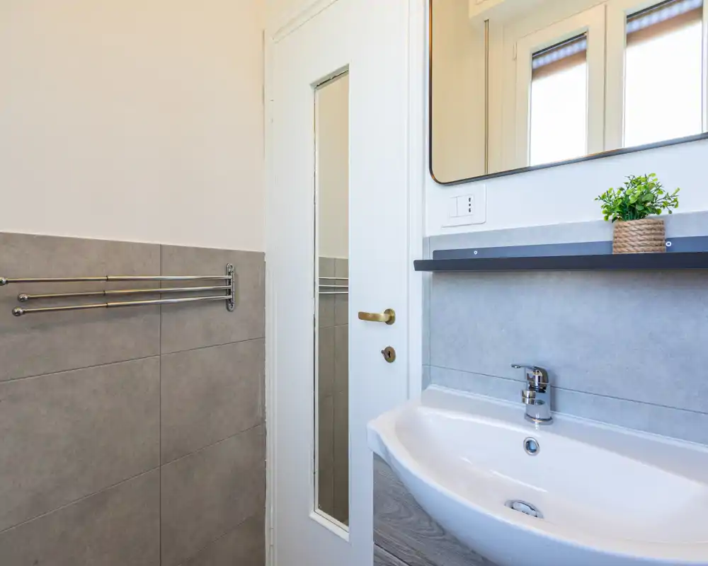Bagno moderno e luminoso con lavabo a parete, rubinetto cromato e grande specchio; piastrelle grigie e una pianta decorativa creano un effetto pulito e fresco.
