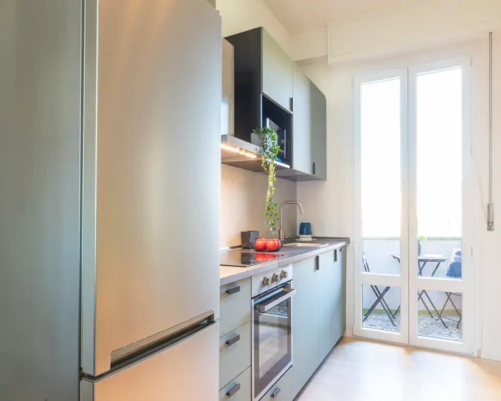 Cucina moderna e luminosa con elettrodomestici integrati, frigorifero in acciaio, forno incassato e piano che si apre su un piccolo balcone — piante e dettagli curati rendono l'ambiente accogliente.