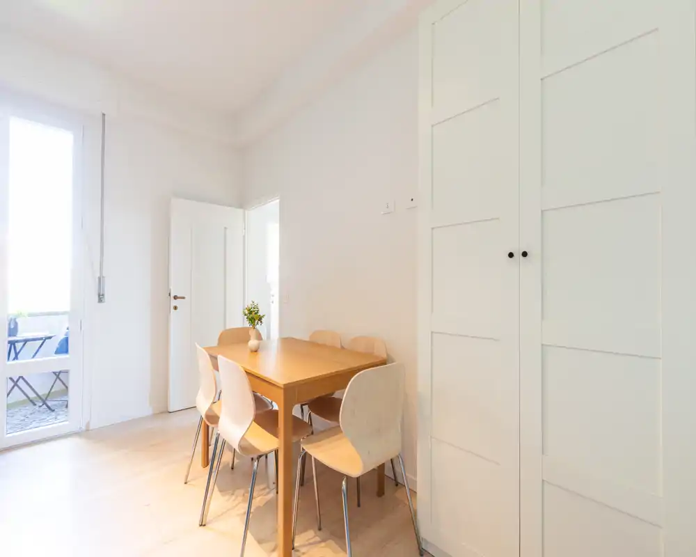 Zona pranzo luminosa e minimal con tavolo in legno per sei persone, sedie chiare e un vaso di fiori. Ampia finestra/porta garantisce luce naturale e un'atmosfera ariosa.