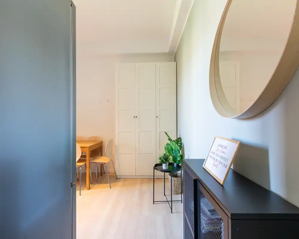 Angolo pranzo compatto con tavolo e sedie in legno accanto a un armadio alto bianco; arredo minimal, luminoso, con pianta decorativa e specchio rotondo.