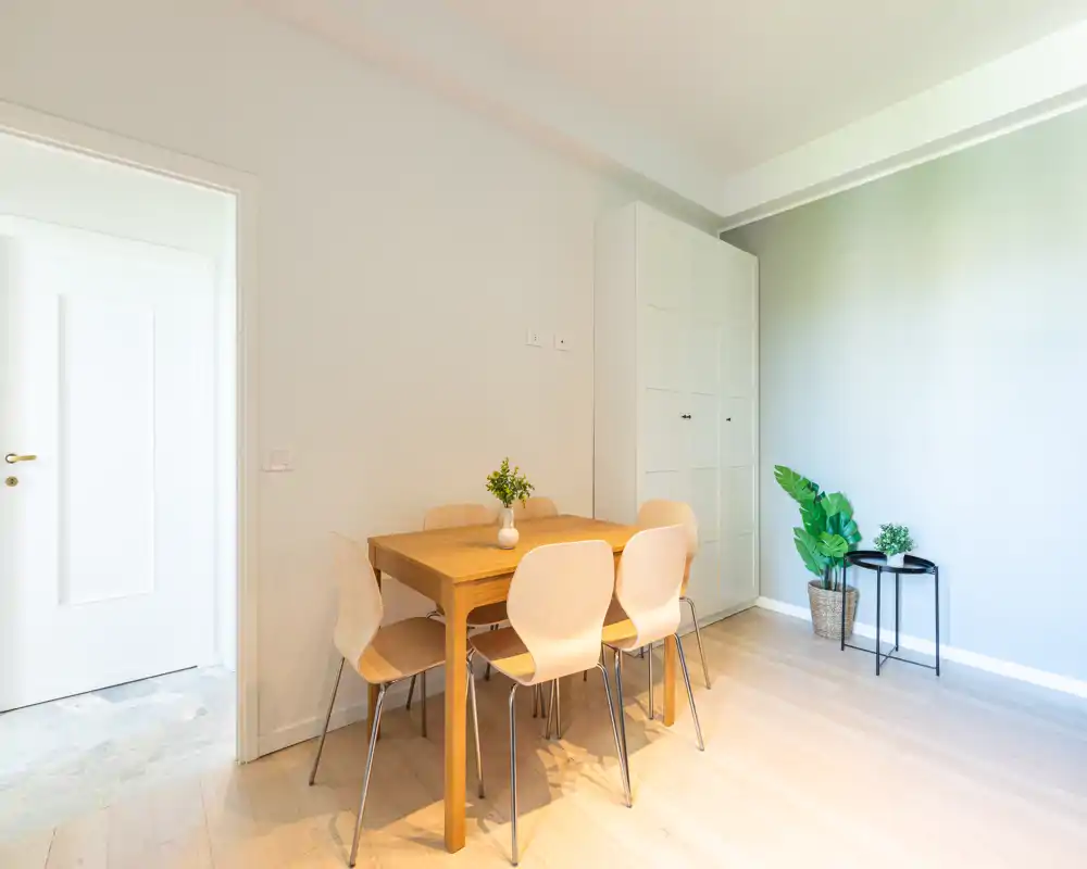 Zona pranzo luminosa e minimal con tavolo in legno e quattro sedie chiare, completata da un armadio alto e piante in vaso per un ambiente accogliente.