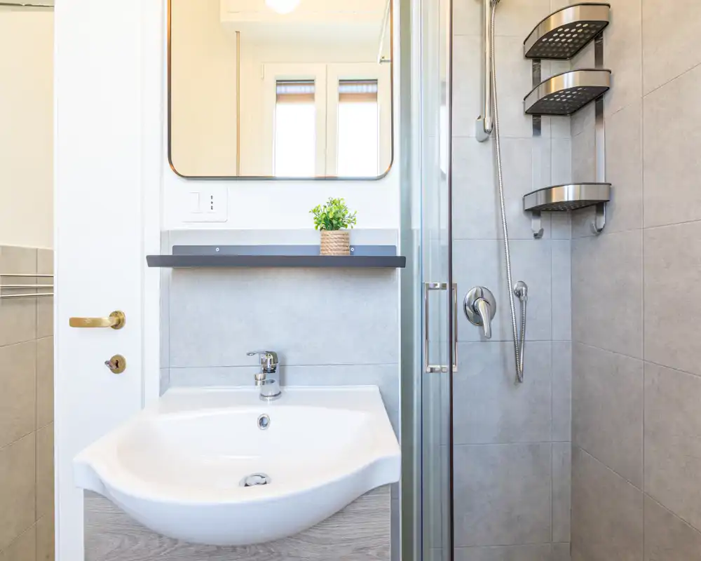 Bagno moderno e compatto con lavabo bianco, rubinetto a parete, specchio e doccia con porta in vetro e accessori in acciaio inox.