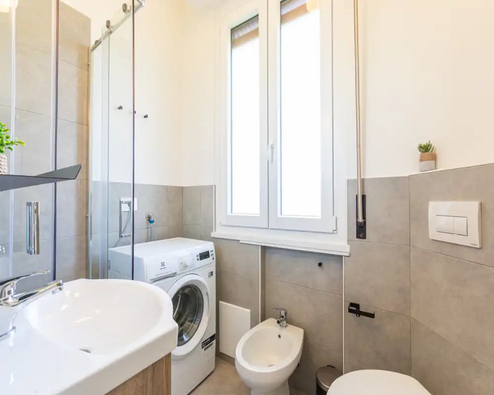 Bagno moderno e luminoso con lavabo, bidet, WC e una lavatrice a carica frontale sotto una finestra ampia. Piastrelle neutre e una cabina doccia in vetro rendono l'ambiente funzionale e curato.