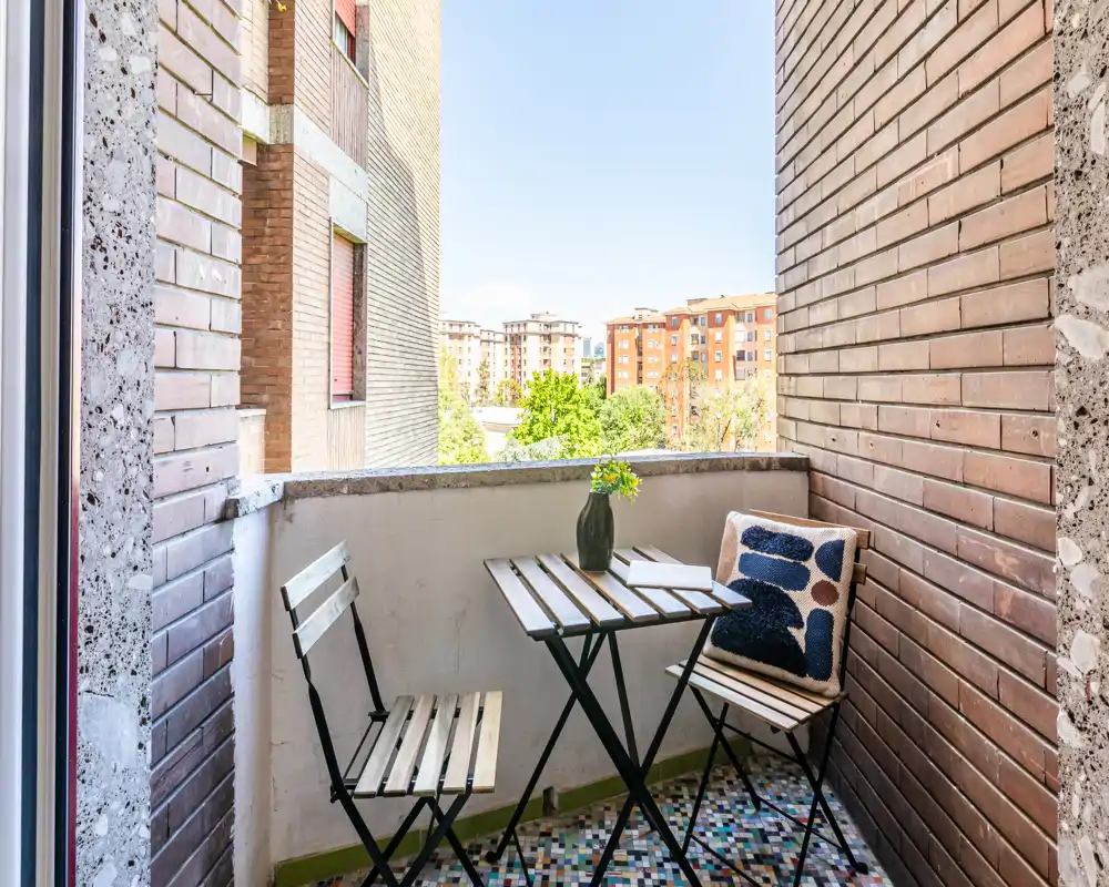 Piccolo balcone accogliente con tavolino bistro pieghevole e due sedie, cuscino decorativo e vaso con fiori; pavimento piastrellato e vista sulla città.