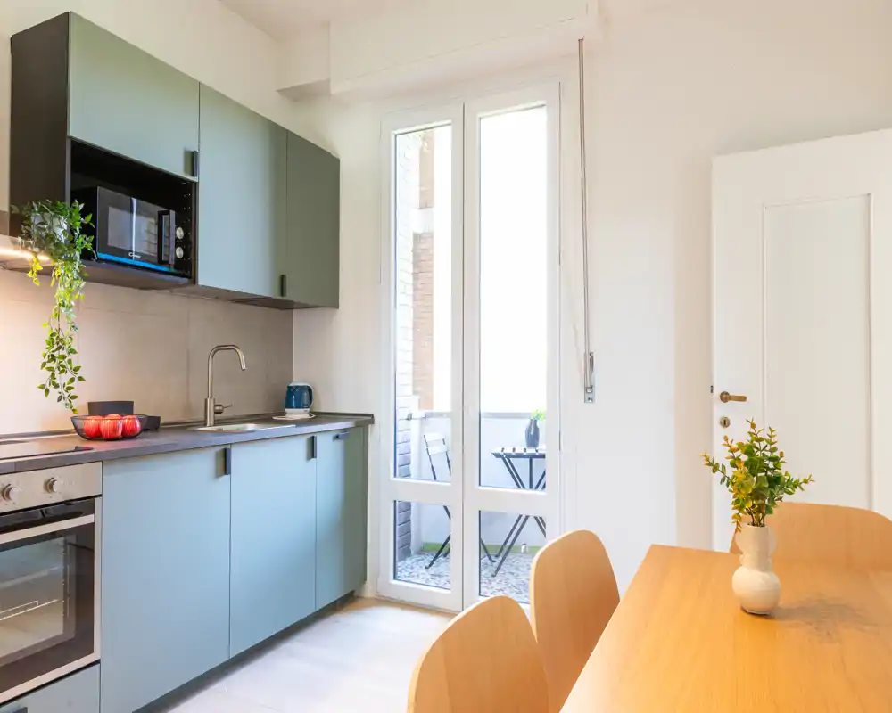 Cucina moderna con mobili azzurri, forno e microonde integrati e un tavolo in legno vicino a una porta finestra luminosa che conduce a un piccolo balcone.