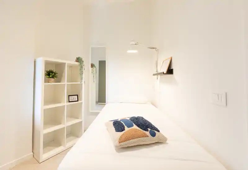 Dormitorio individual pequeño y luminoso con cama bien hecha, cojín decorativo, lámpara de pared y estantería — estilo minimalista y limpio, ideal para un huésped.