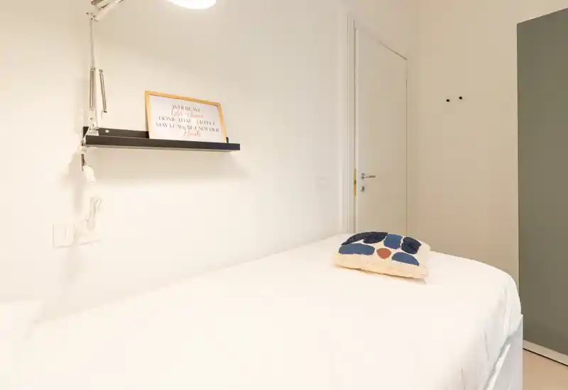 Dormitorio individual luminoso y minimalista con cama bien hecha, cojín decorativo y estantería de pared; decoración neutra y limpia, ideal para un alquiler compacto.